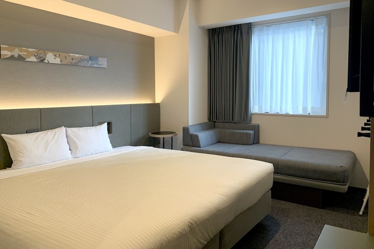 A hotel room with a large bed, a smaller sofa, a bedside table, a window with curtains, and a wall-mounted artwork.
