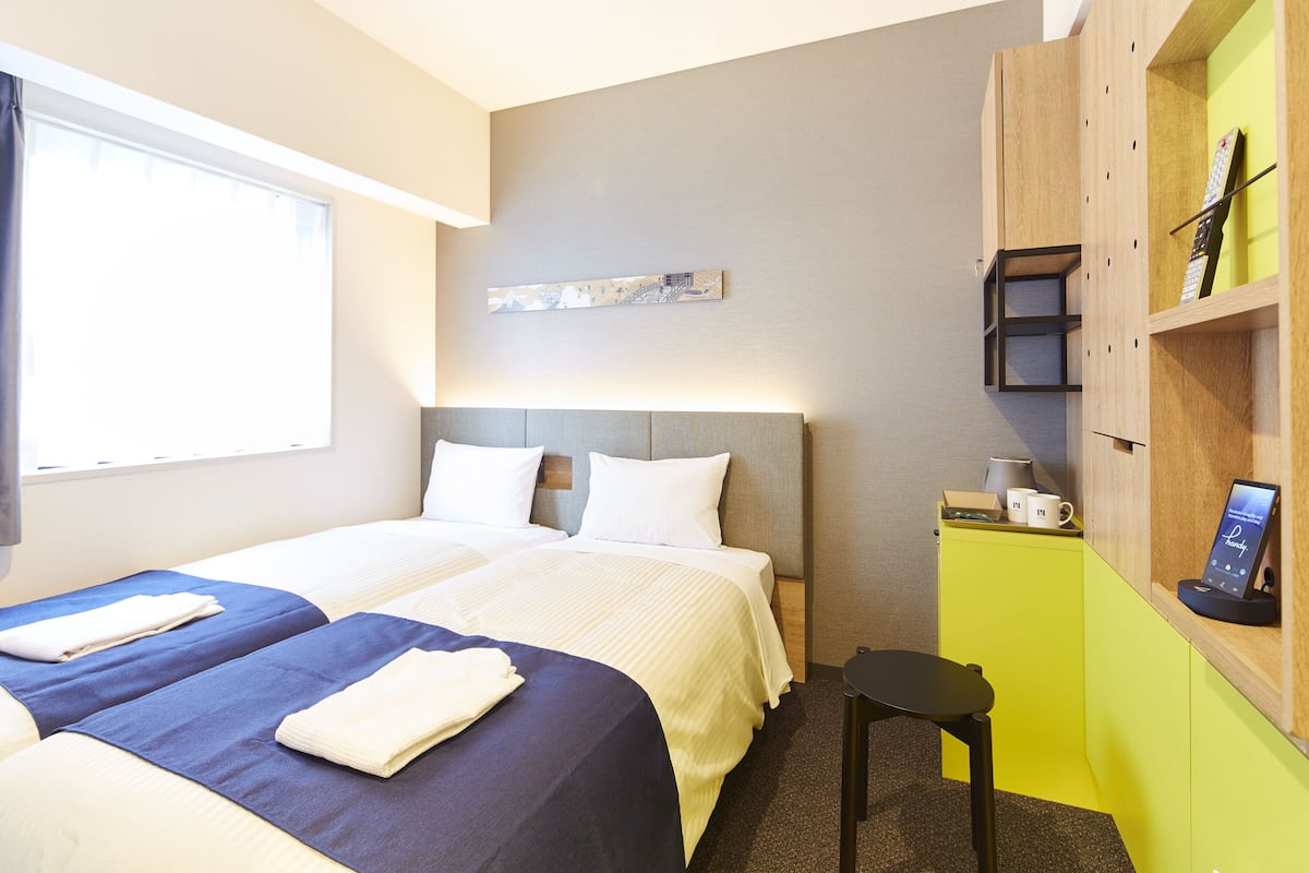 A hotel room with two beds, a desk, a chair, a small table with a coffee cup, and a wall-mounted shelf with a clock and a framed picture.
