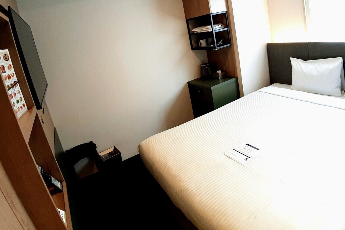 A hotel room with a bed, a small table, a shelf with towels, and a wall-mounted TV.