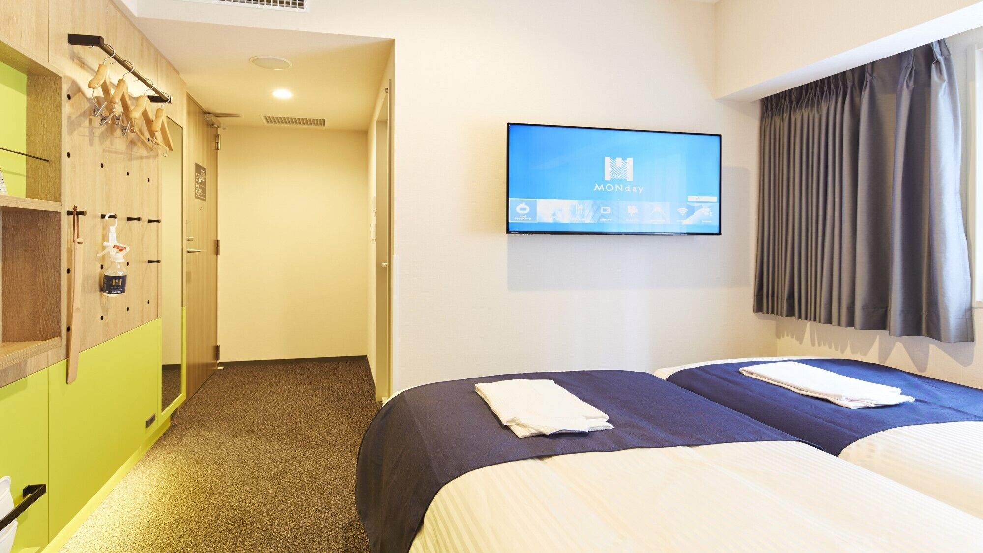 Quarto de hotel com duas camas, uma televisão na parede, um cabideiro e um guarda-roupa de madeira.
