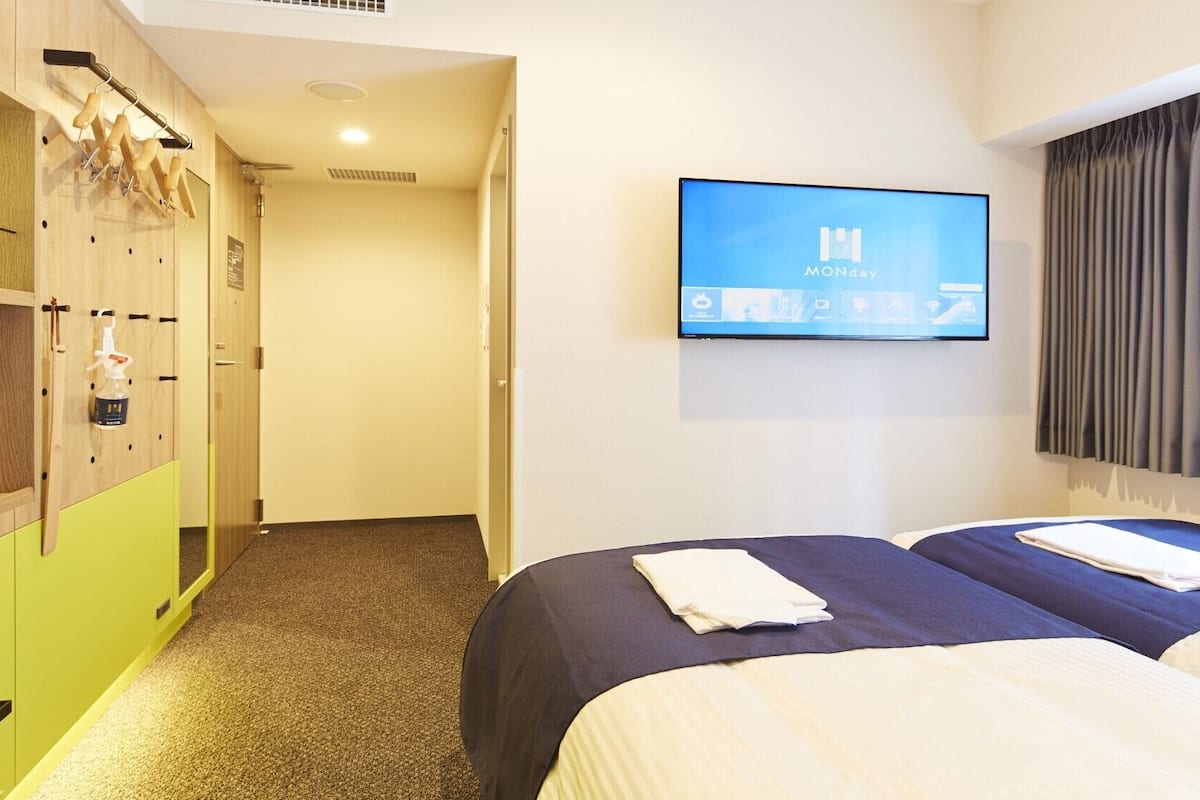 A hotel room with two beds, a TV on the wall, a coat rack, and a wooden wardrobe.