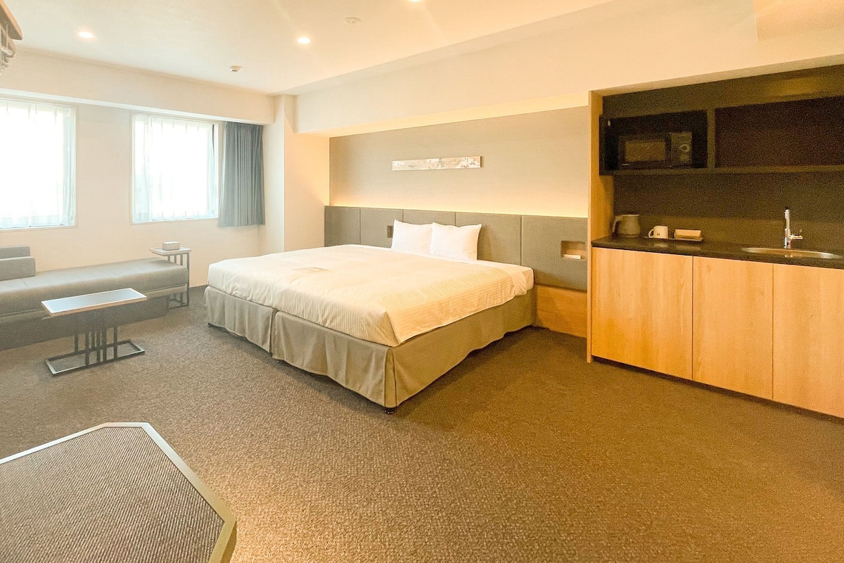 A hotel room with a bed, a seating area, a kitchenette, and a large window.