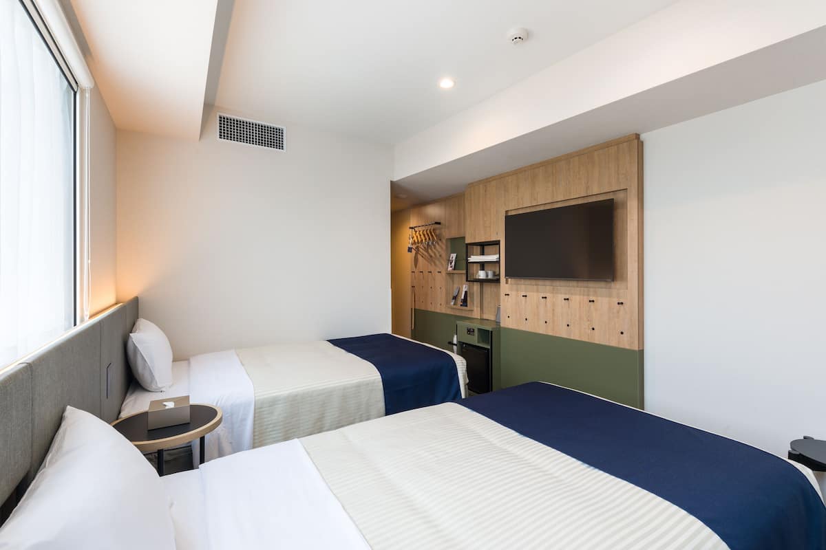 A hotel room with two beds, a small table, a desk, and a wardrobe with hanging clothes.