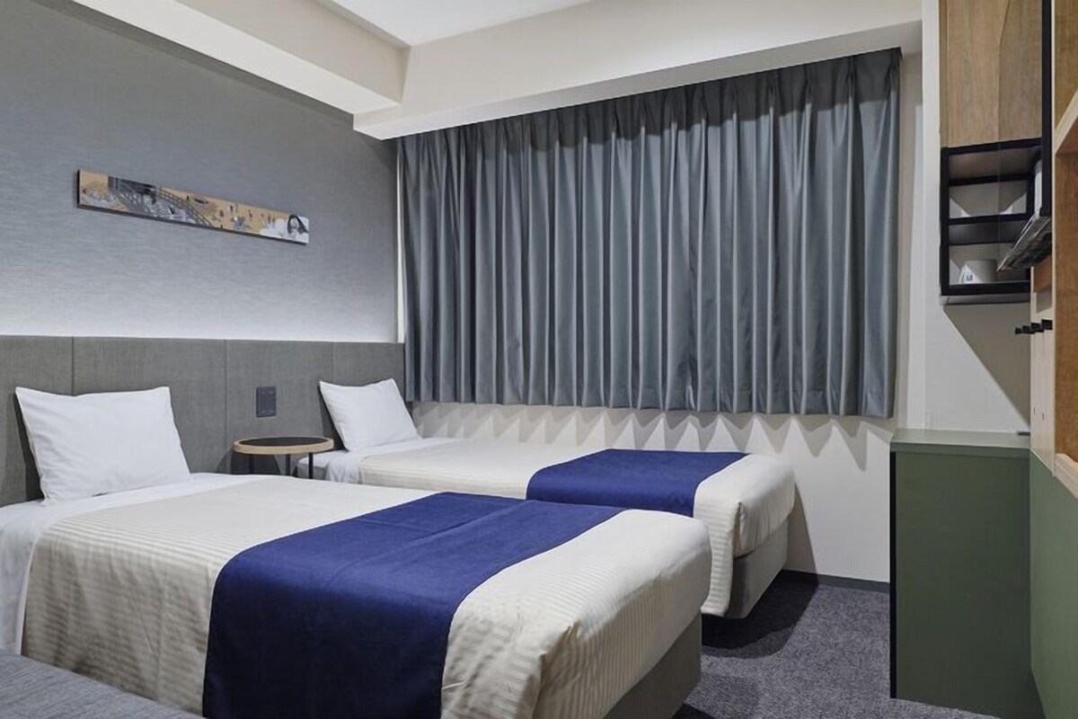 A hotel room with two beds, a small table, a painting on the wall, and a window with curtains.