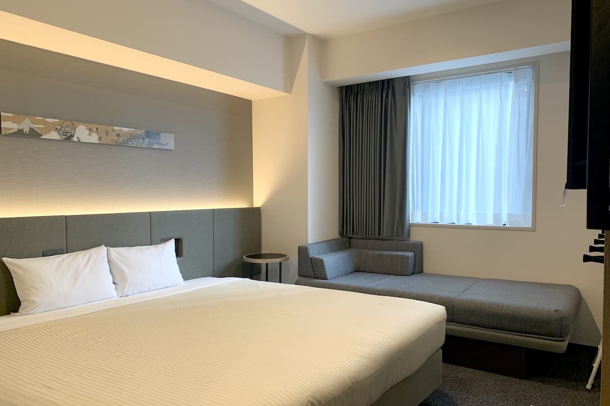 A hotel room with a large bed, a smaller sofa, a side table, and a window with curtains.