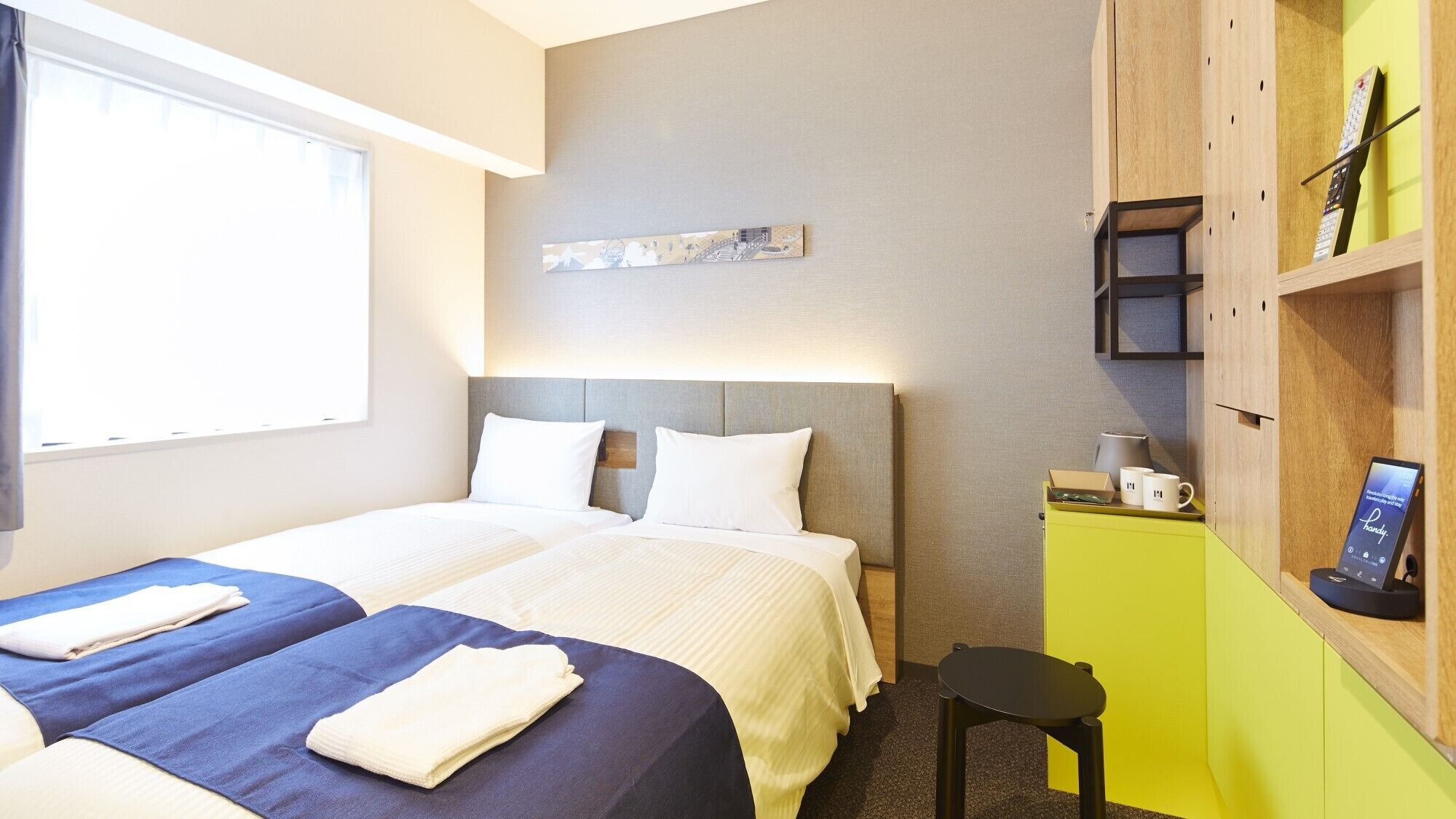 A hotel room with two beds, a desk, a chair, a shelf with a remote control, and a wall-mounted artwork.