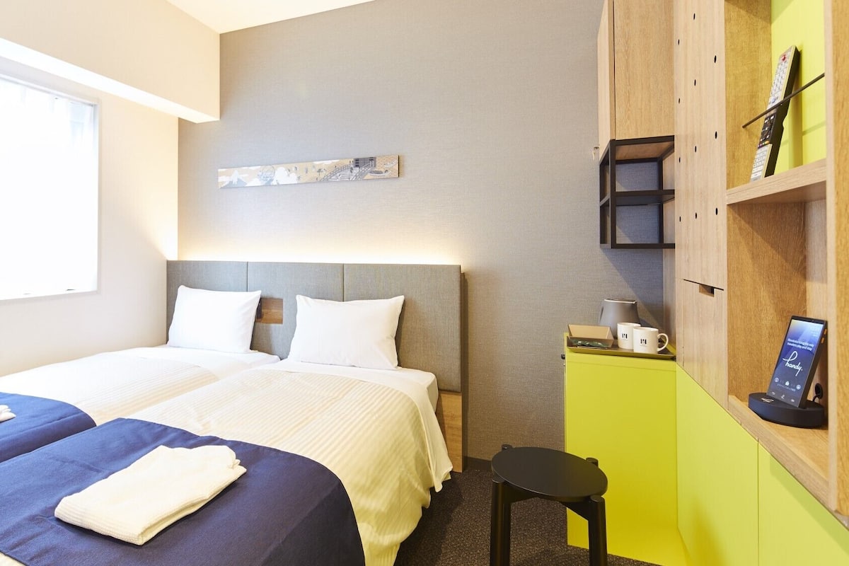 A hotel room with two beds, a desk, a chair, a shelf with a remote control, and a wall-mounted artwork.