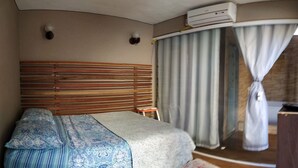 Quarto de hotel com cama, uma mesinha, ar condicionado e janela com cortinas.