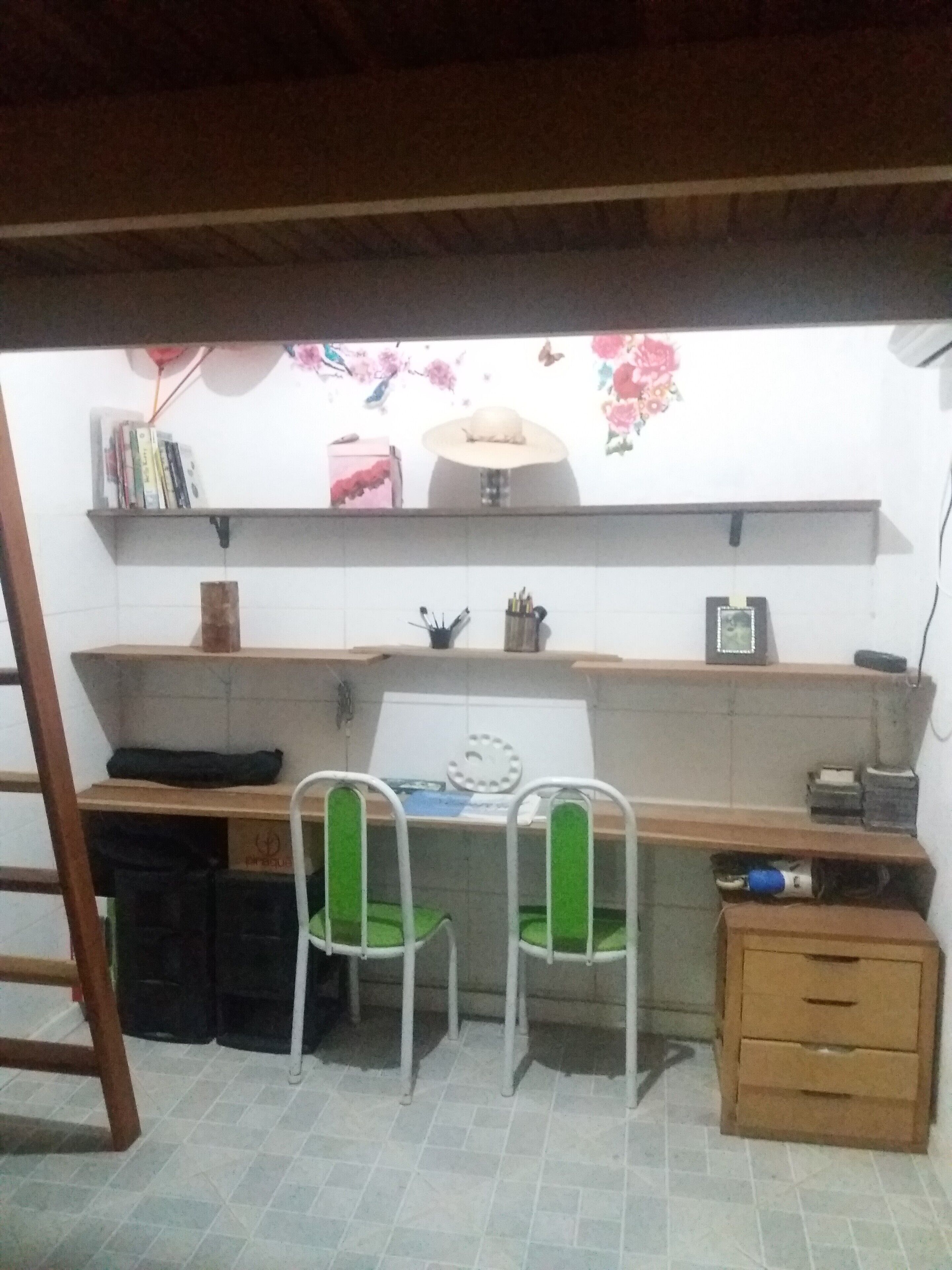 Uma cozinha pequena e compacta com painel de azulejos, uma pia e bancada. Há duas cadeiras com almofadas verdes, um armário de madeira e uma escada encostada na parede.