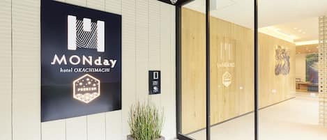 A hotel entrance with a large sign reading "MONday hotel OKACHIMACHI PREMIUM" and a reception area visible through glass doors.