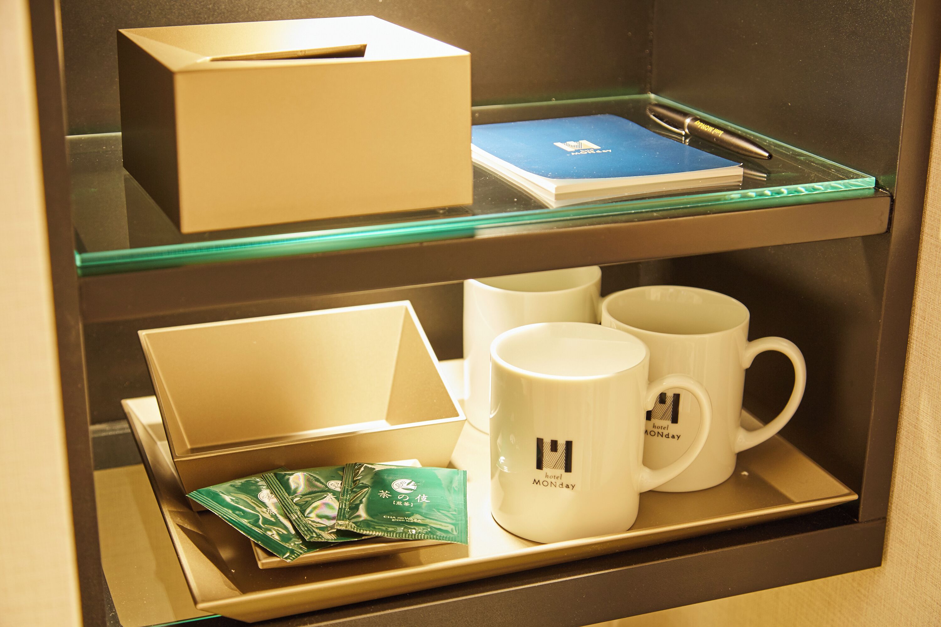 A hotel minibar with a glass shelf containing a box, a book, two mugs, and a packet of tea.
