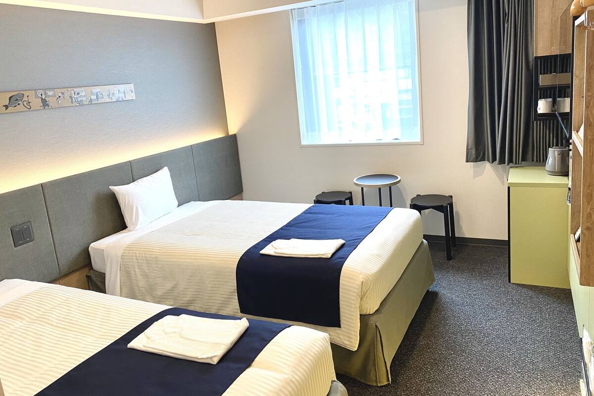 A hotel room with two beds, a small table, a chair, a window with curtains, and a kitchenette.