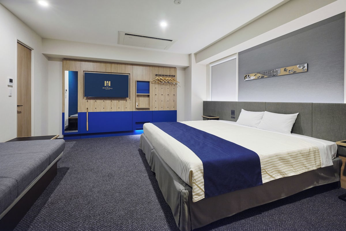A modern hotel room with a large bed, a smaller bed, a wardrobe, a seating area, and a television.