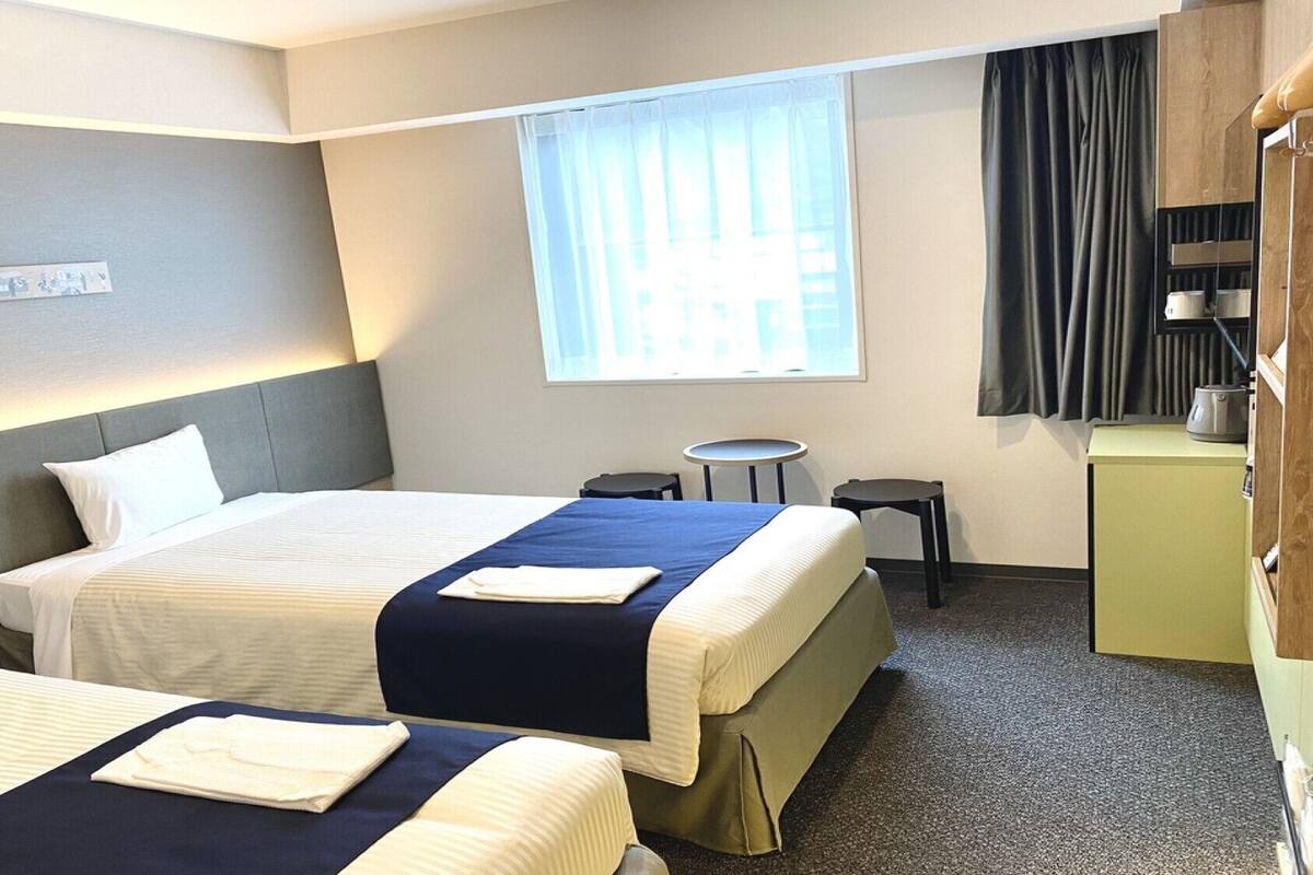 A hotel room with two beds, a small table, a chair, a window with curtains, and a kitchenette area.