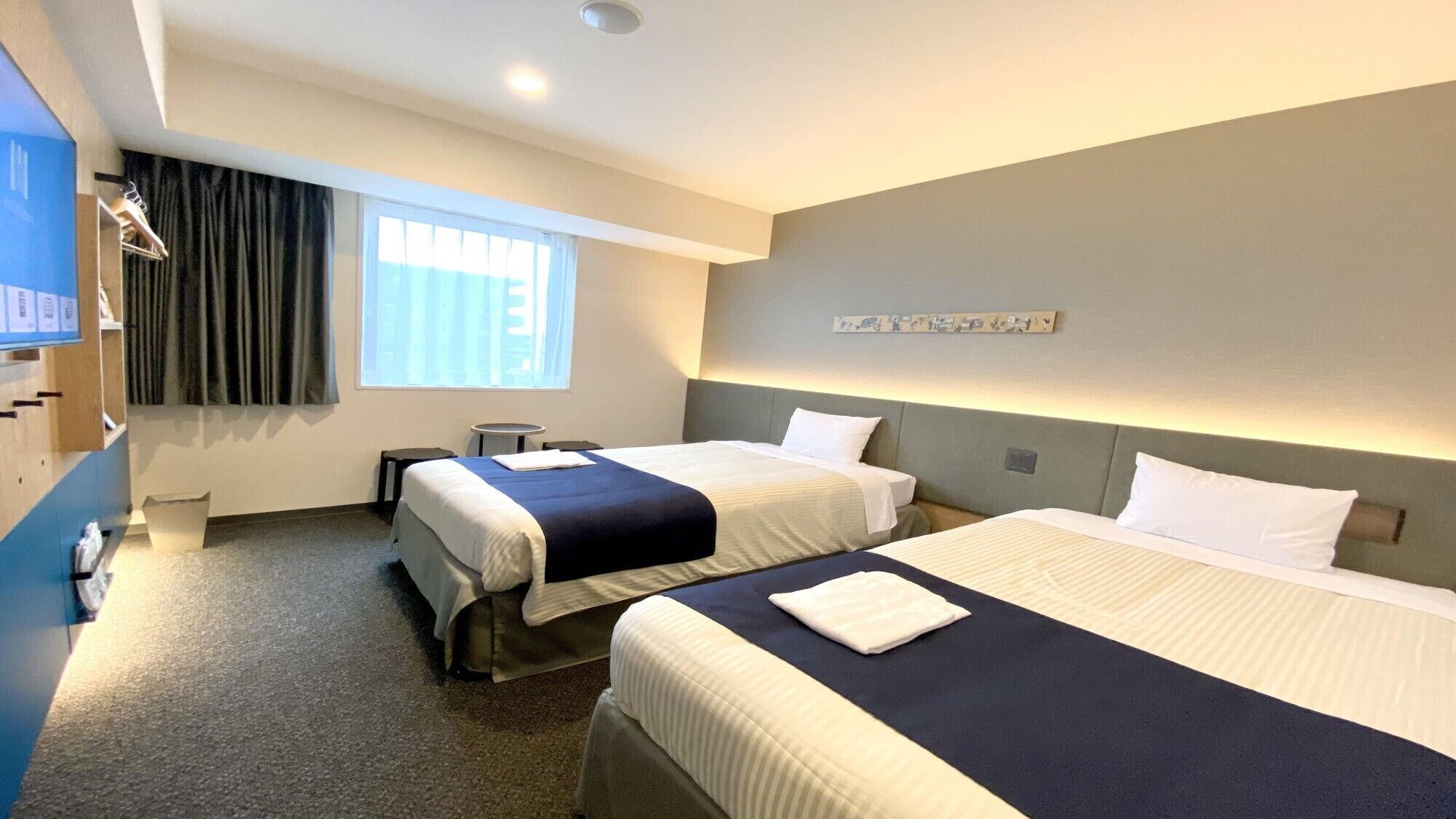 A hotel room with two beds, a desk, a chair, a small table, a window with curtains, and a wall-mounted artwork.