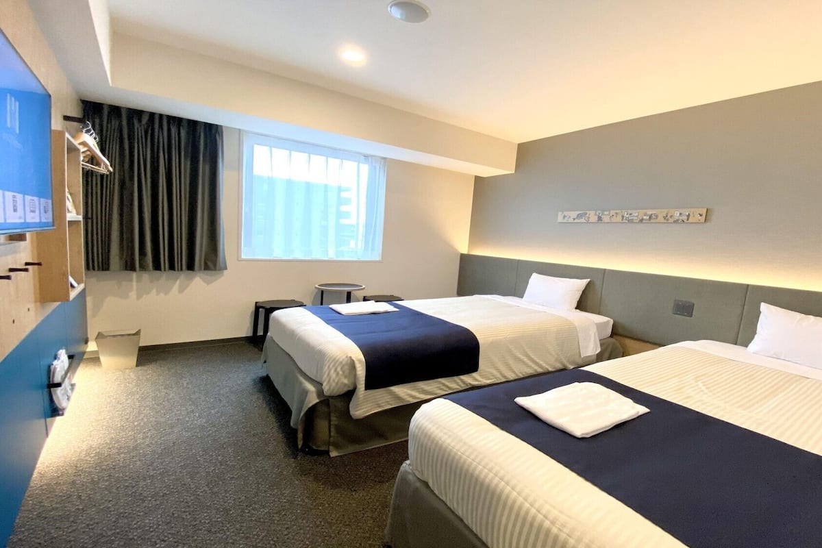 A hotel room with two beds, a desk, a chair, a small table, a window with curtains, and a wall-mounted artwork.