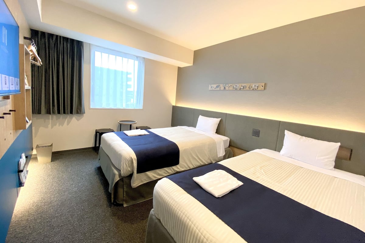 A hotel room with two beds, a small table, a chair, a window with curtains, a wall-mounted artwork, and a ceiling light.