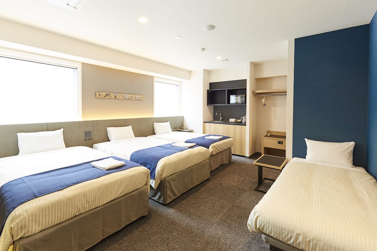 A hotel room with three beds, a small kitchenette, and a large window.