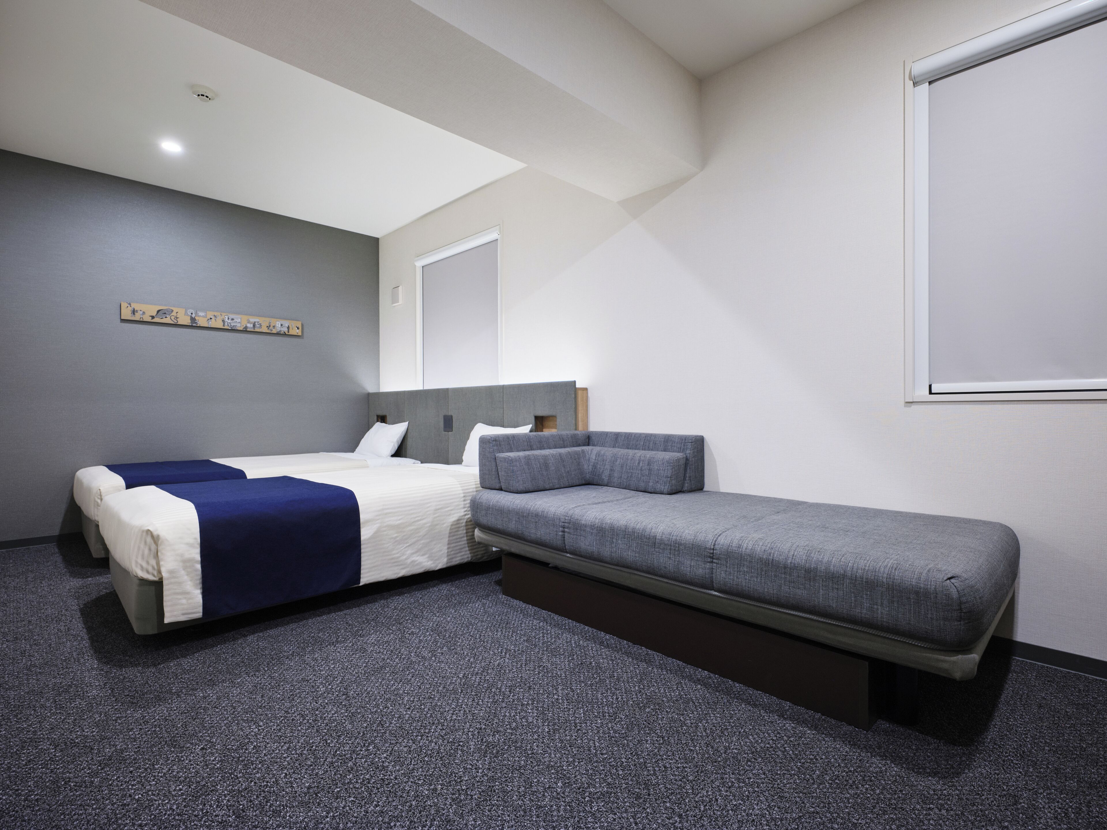 A modern hotel room with a bed, a sofa, a carpeted floor, and a wall-mounted projector screen.