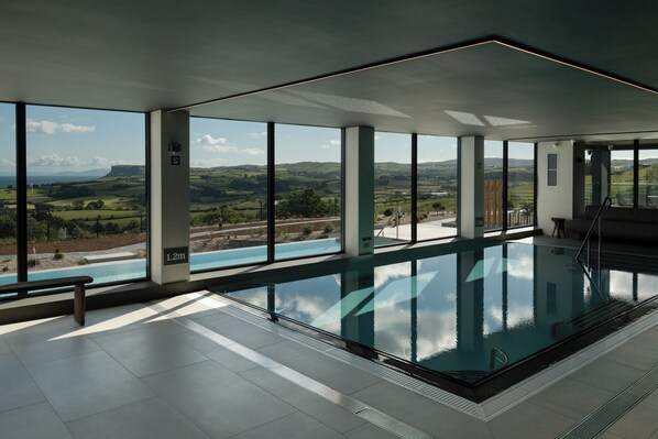 A modern indoor swimming pool with large windows offering a view of a green landscape.