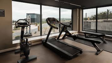 Un gimnasio con cintas de correr y una máquina elíptica, amplias ventanas con vistas a un edificio y un paisaje, y suelo de madera.