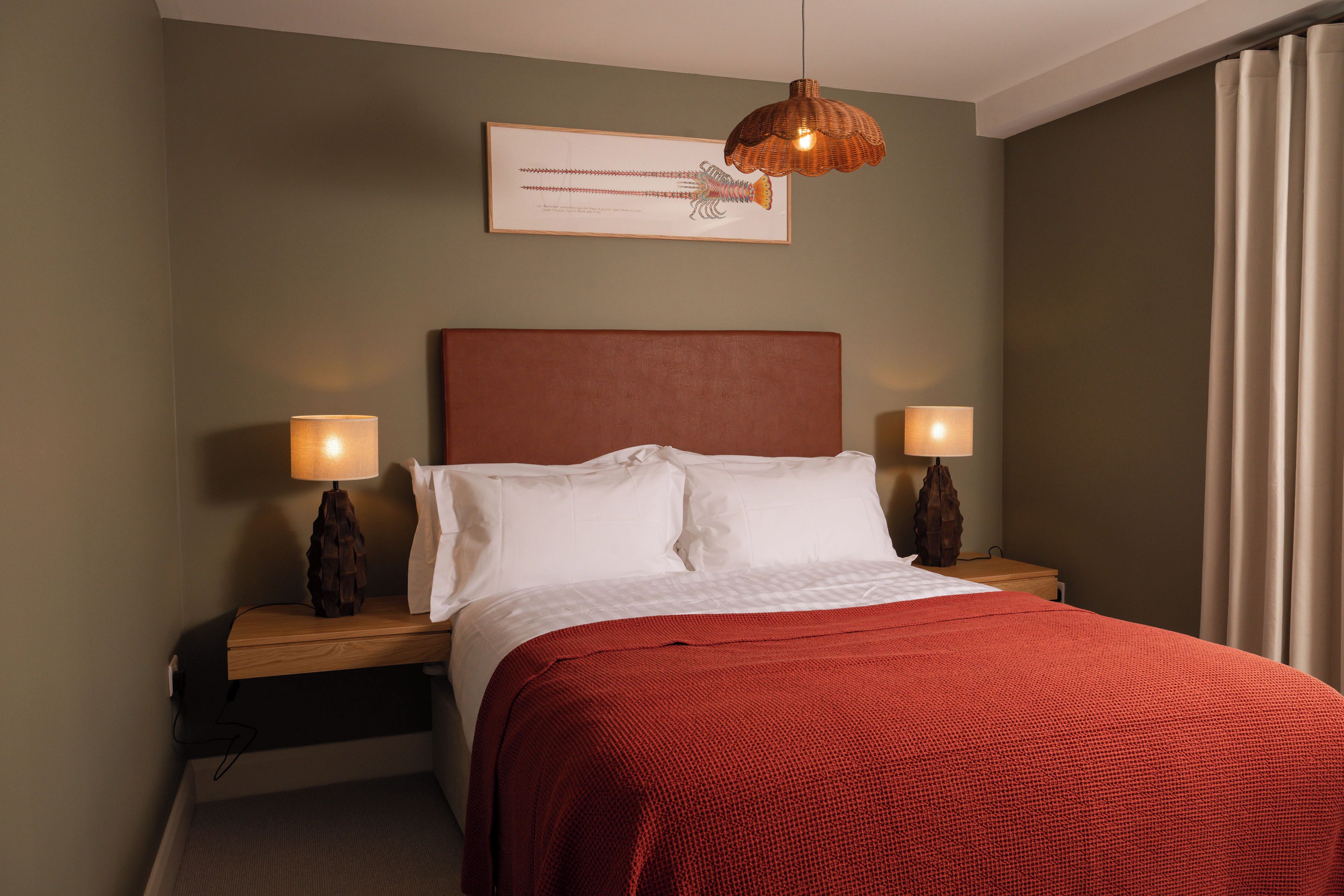 A neatly made bed with a red blanket, two bedside lamps, and a framed artwork on the wall.