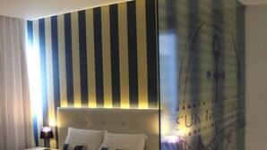 A hotel room with a bed, a nightstand, a lamp, a glass partition, and a wall with striped wallpaper. - Costa de Prata Hotel (Figueira da Foz)