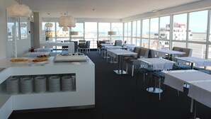 A modern restaurant with a buffet area, dining tables, and large windows offering a city view. - Costa de Prata Hotel (Figueira da Foz)