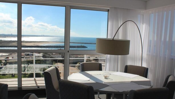 A dining area with a table and chairs, large windows with a view of the sea and a distant shoreline, and a floor lamp. - Costa de Prata Hotel (Figueira da Foz)