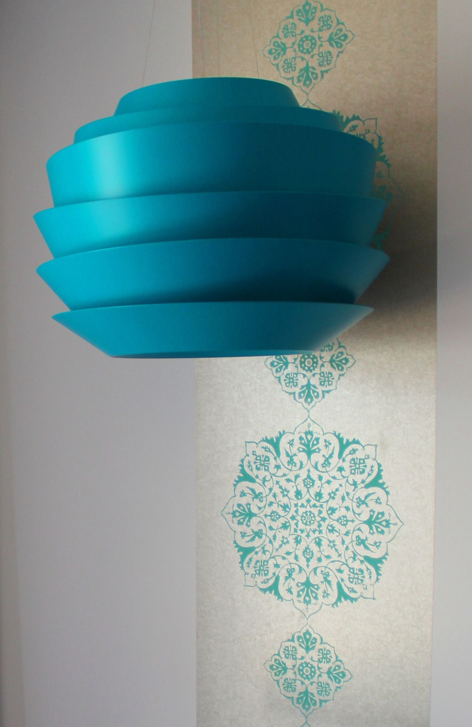 A teal-colored, multi-tiered hanging light fixture against a wall with a decorative, geometric pattern.