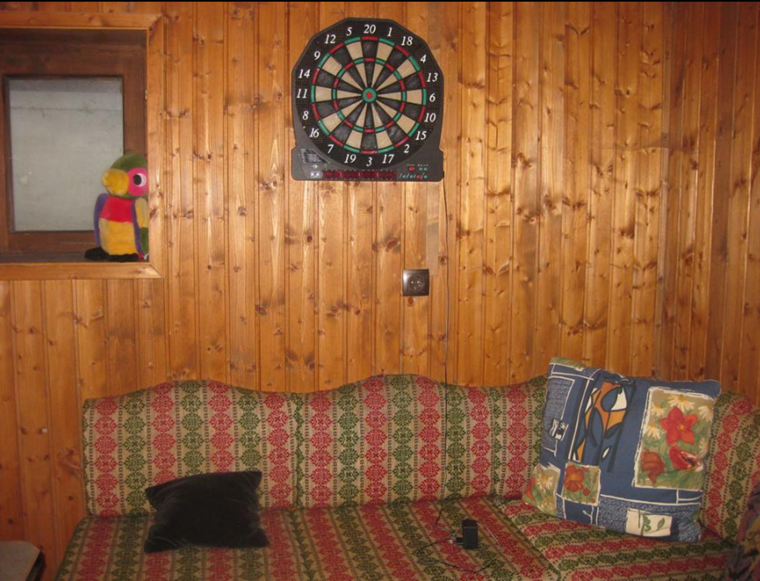 Ein Zimmer mit Dartscheibe, einem farbenfrohen gemusterten Sofa und einem Stofftier auf einem Regal.