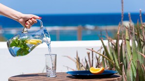 A hand pouring water from a pitcher into a glass on a wooden table with a blue plate and an orange slice. - Domus Litus (Milos)