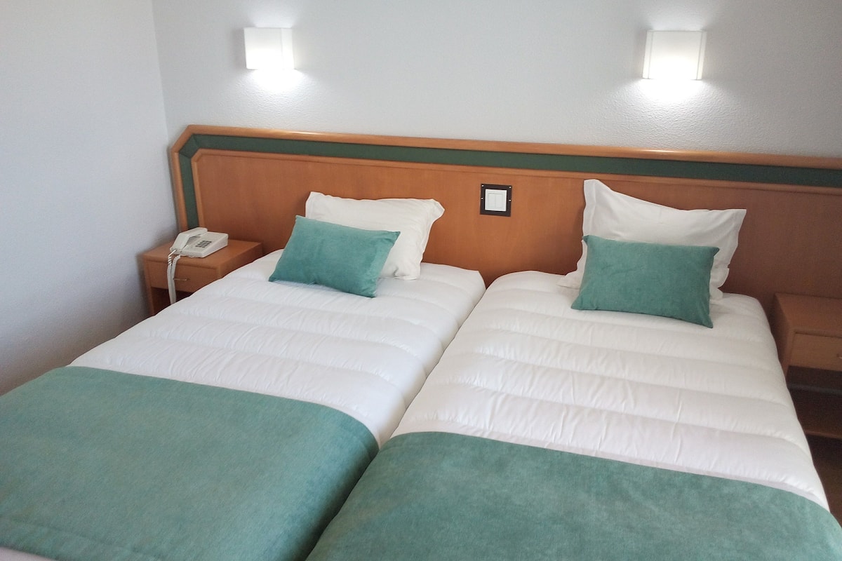 Habitación de hotel con dos camas individuales, cada una con almohada verde y ropa de cama blanca. Se observa un cabecero de madera, una mesita de noche con teléfono y una lámpara de pared.