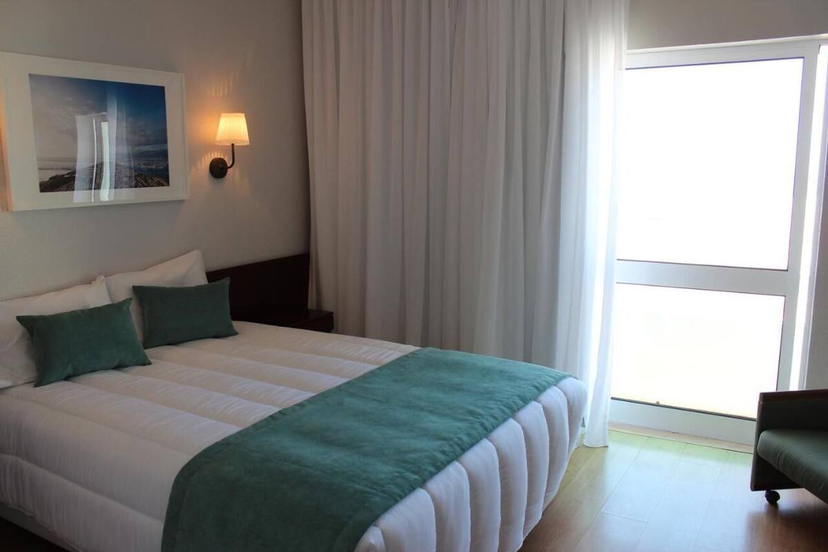 Habitación de hotel con una cama grande, una mesita de noche, una lámpara de pared, una ventana con cortinas y un cuadro enmarcado.