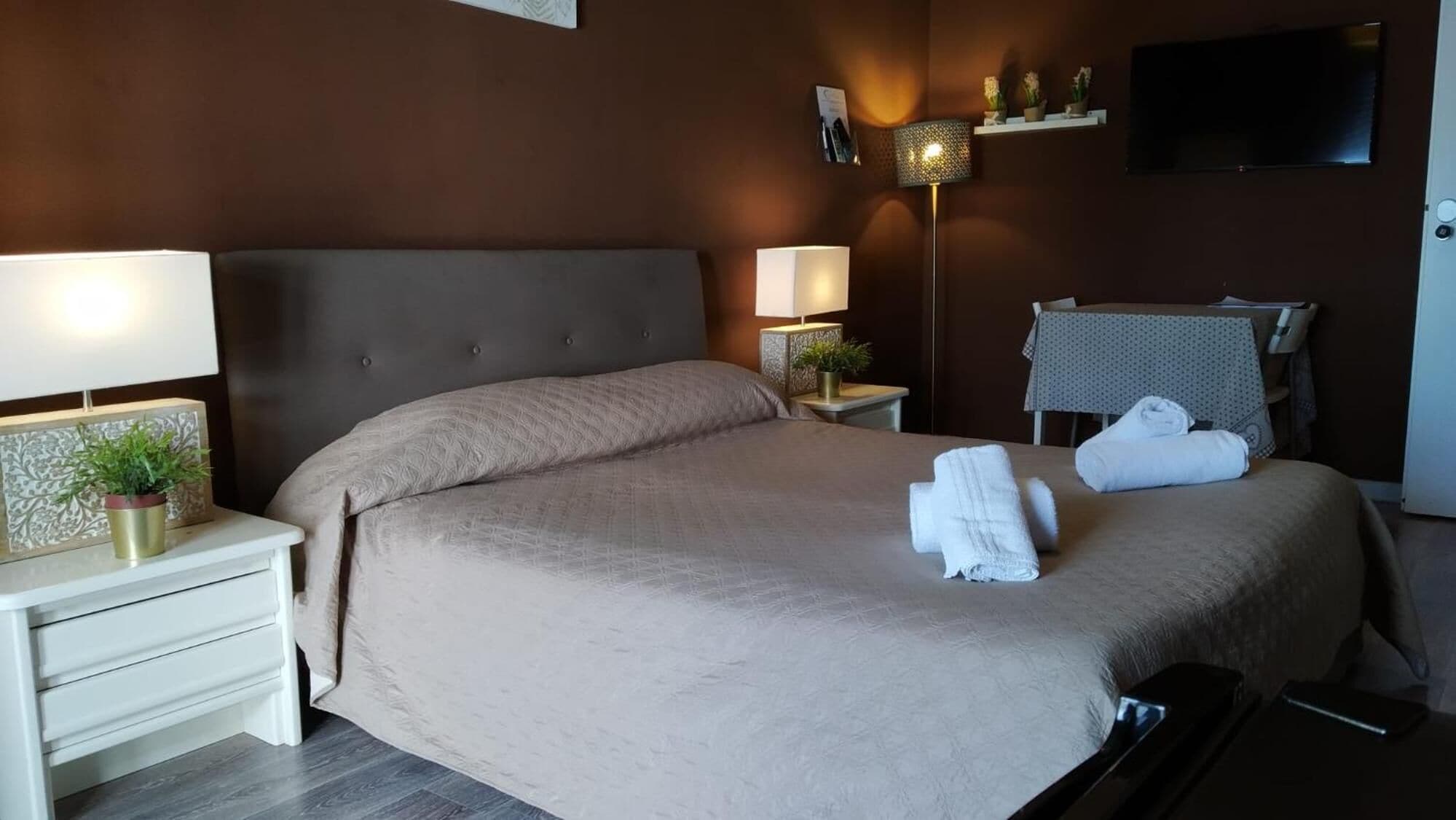 A neatly made bed with a beige bedspread, two bedside lamps, a small potted plant, and a towel on the bed.