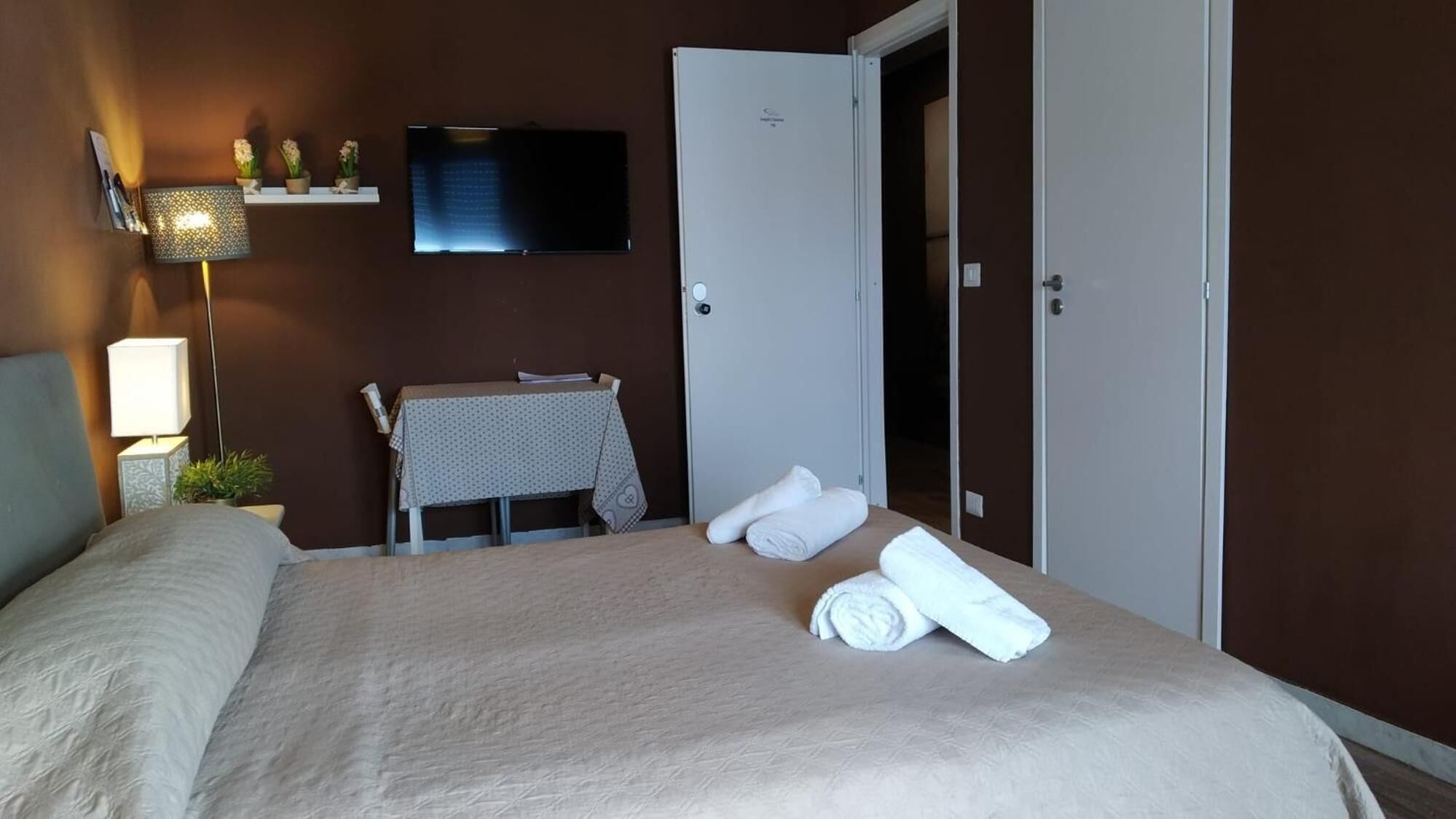 A hotel room with a bed, a table with a tablecloth, a television mounted on the wall, and a door.