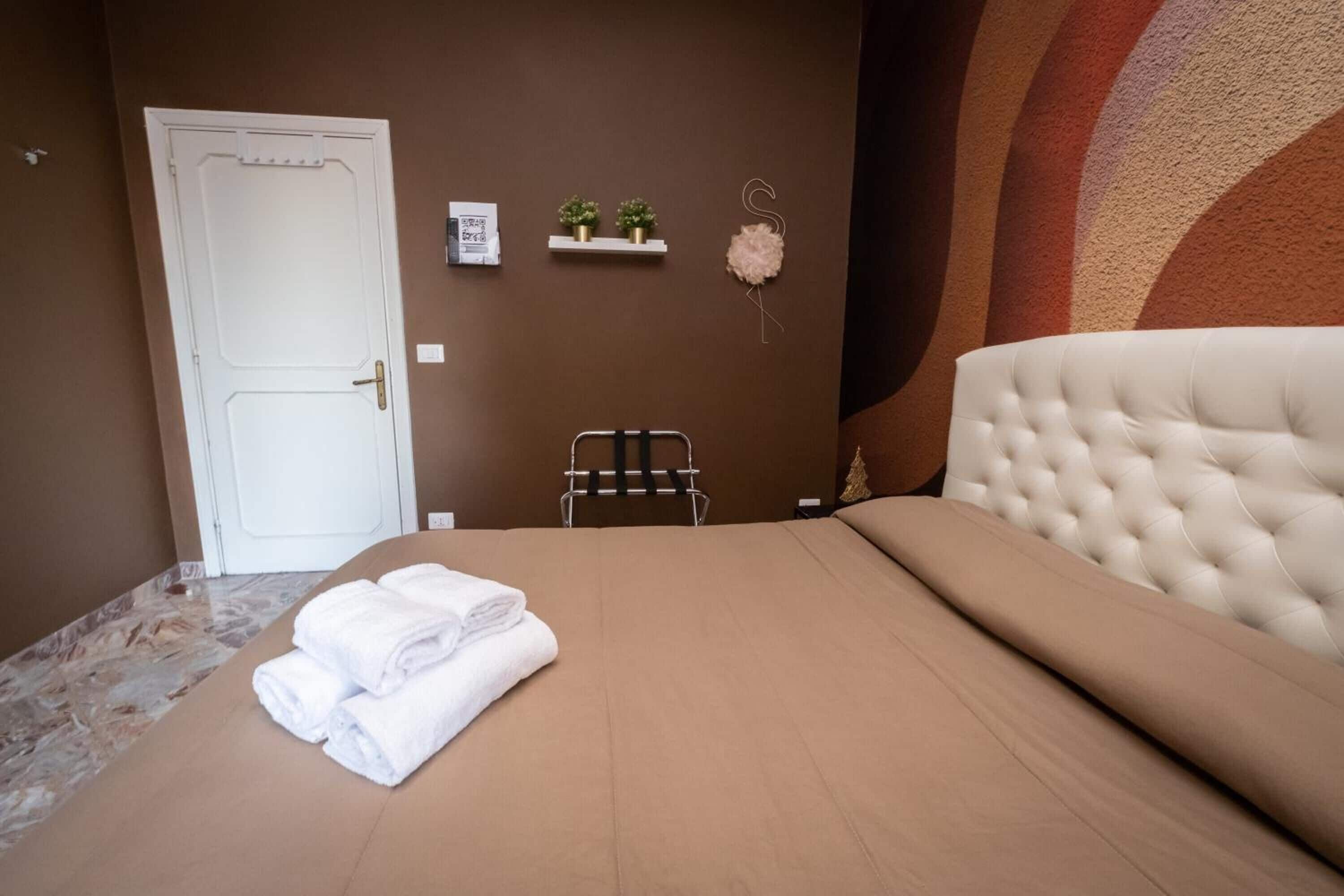 A hotel room with a bed, a white door, a towel rack, a small shelf with plants, and a decorative wall piece.