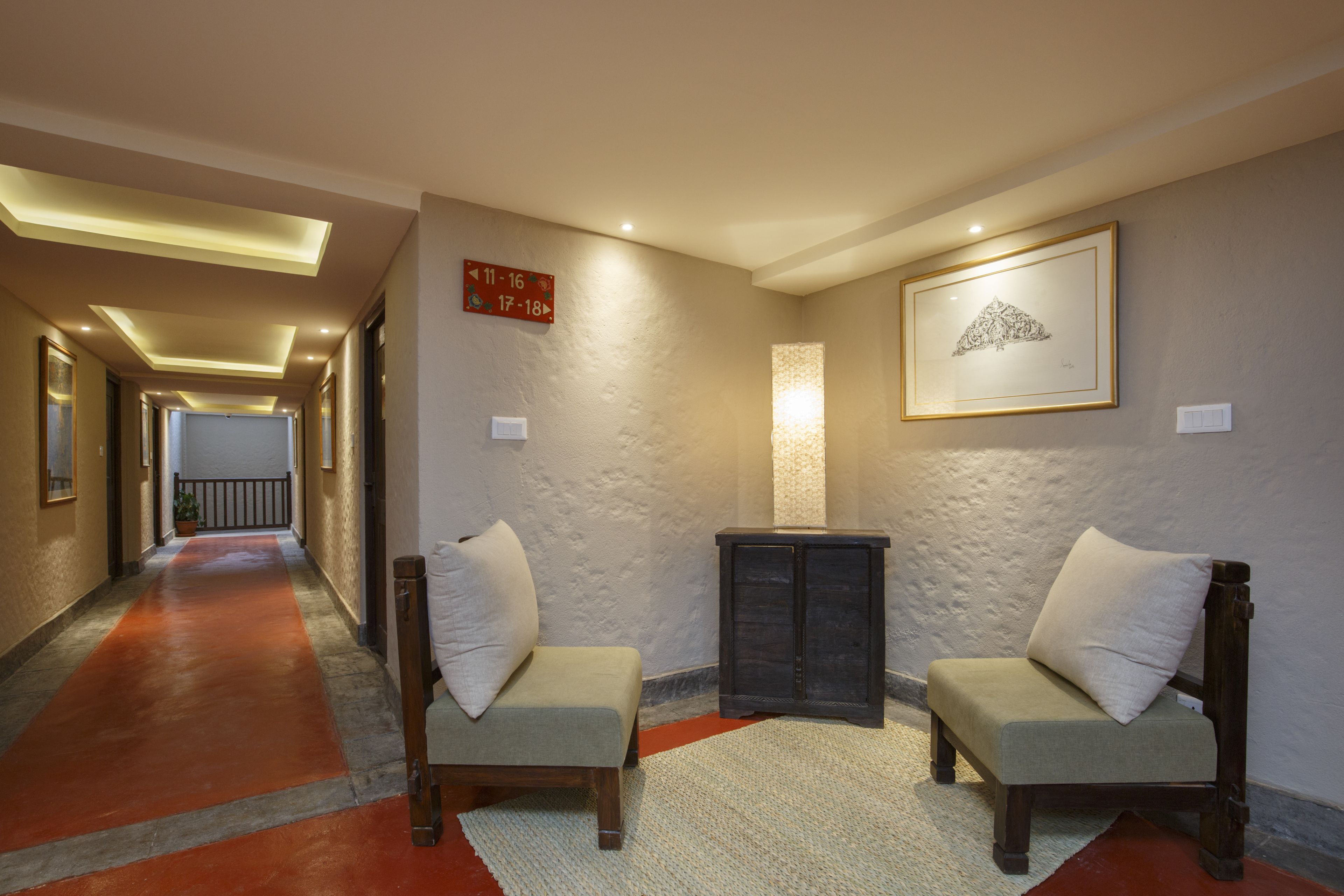 A hallway with a red carpet leading to a reception area. There are two armchairs with cushions, a small cabinet, and a framed artwork on the wall.