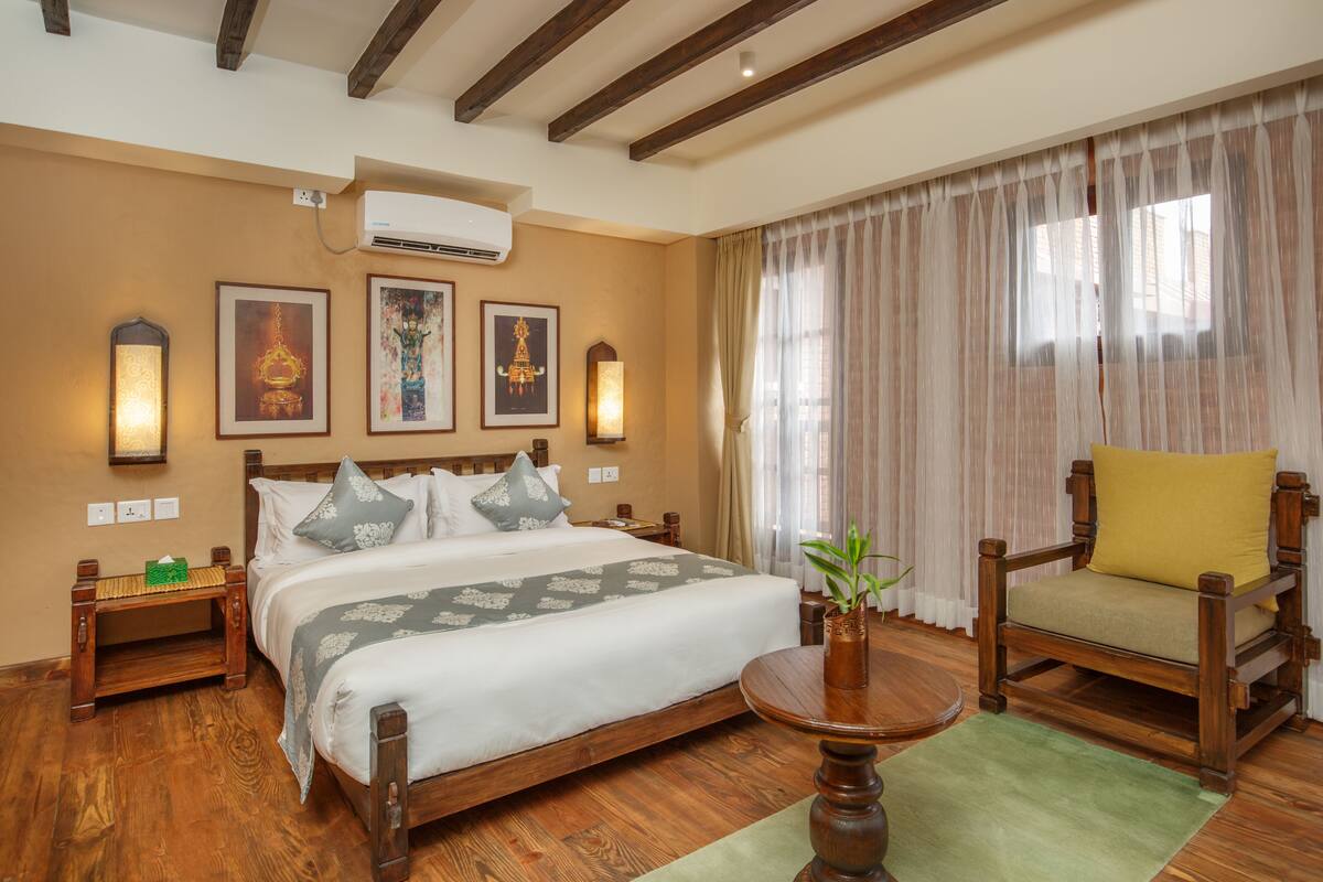A hotel room with a wooden bed, a green rug, a wooden chair with a yellow cushion, and a wall-mounted air conditioner.