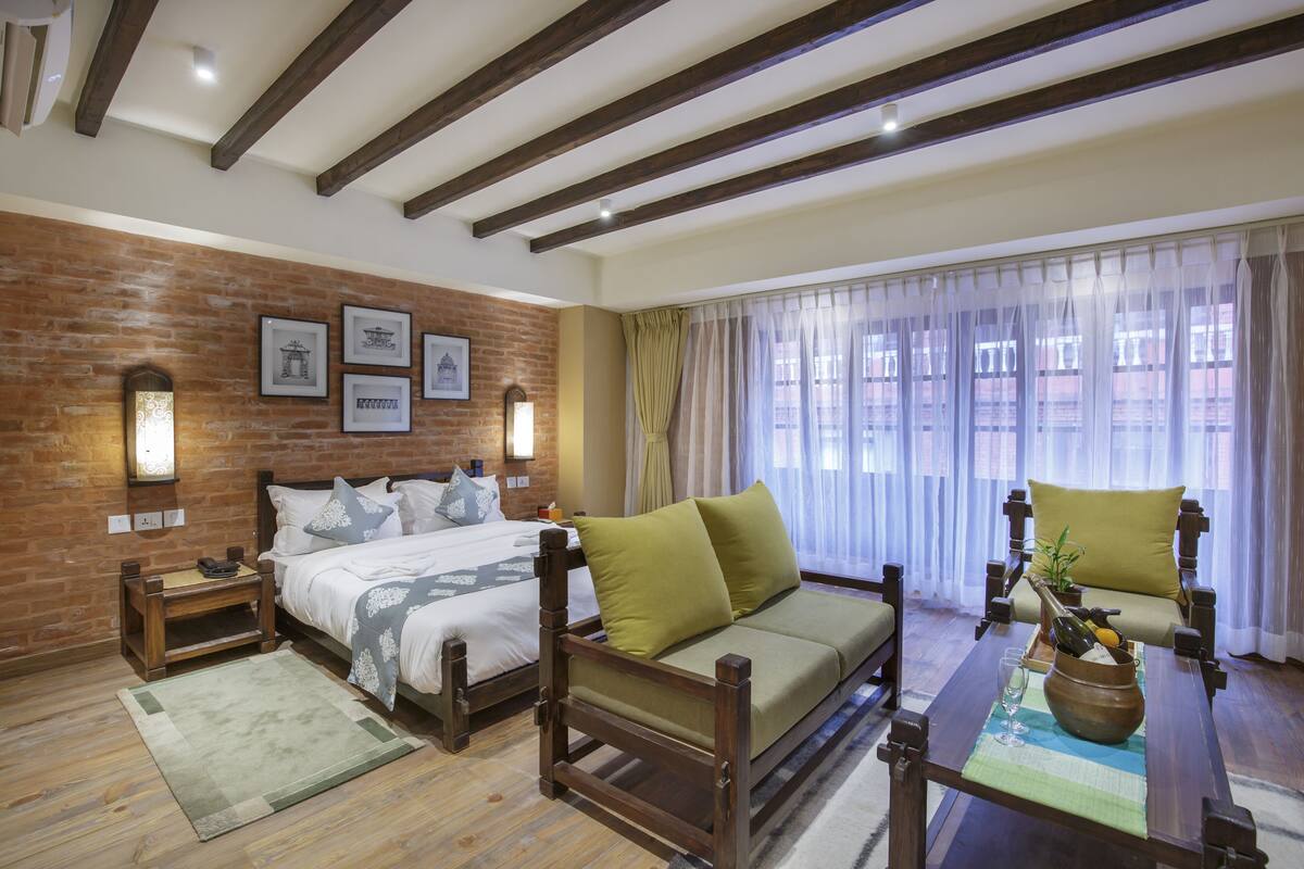 A hotel room with a bed, a sofa, a wooden table, and a brick wall with framed pictures.