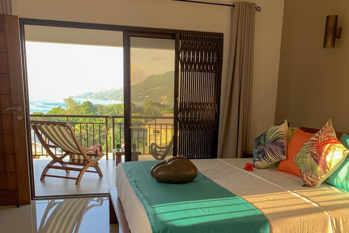 A hotel room with a bed, a view of the sea, a balcony with a chair, and a wooden floor.