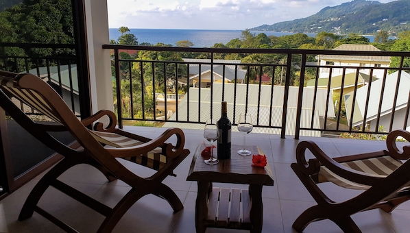 A balcony with two wooden chairs and a table, overlooking a coastal landscape with houses and greenery.
