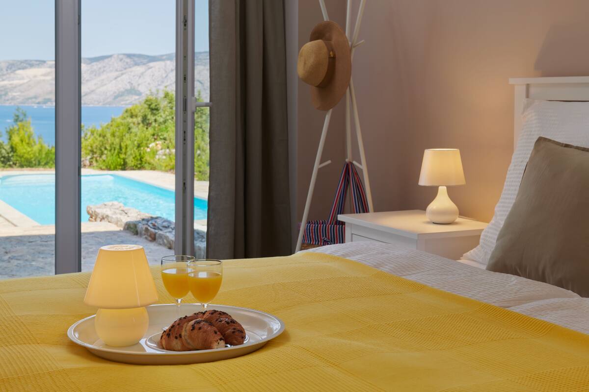 A hotel room with a bed, a bedside table with a lamp, a plate with croissants and orange juice, and a view of a pool and mountains.