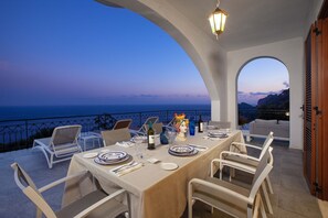Area pranzo all'aperto con vista sul mare, sedie bianche, tavolo apparecchiato per due e una statua decorativa.