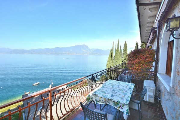 A balcony with a table and chairs, overlooking a lake with boats and mountains in the distance.