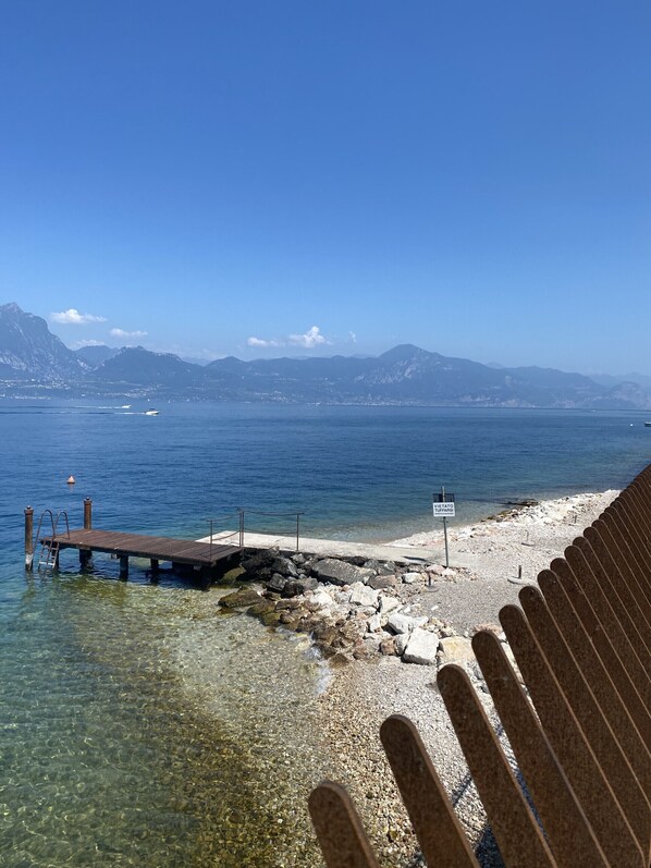A clear view of a rocky shoreline with a wooden pier extending into calm waters, a sign indicating 'Vietato Tuffarsi,' and distant mountains under a clear sky. - Leonardo Walsh Apartment With Lake View (Torri del Benaco)