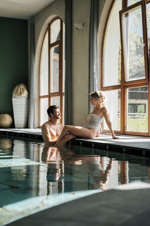 A man and a woman sitting by a poolside window with arched windows and a view of greenery outside. - Hotel La Majun (Badia)