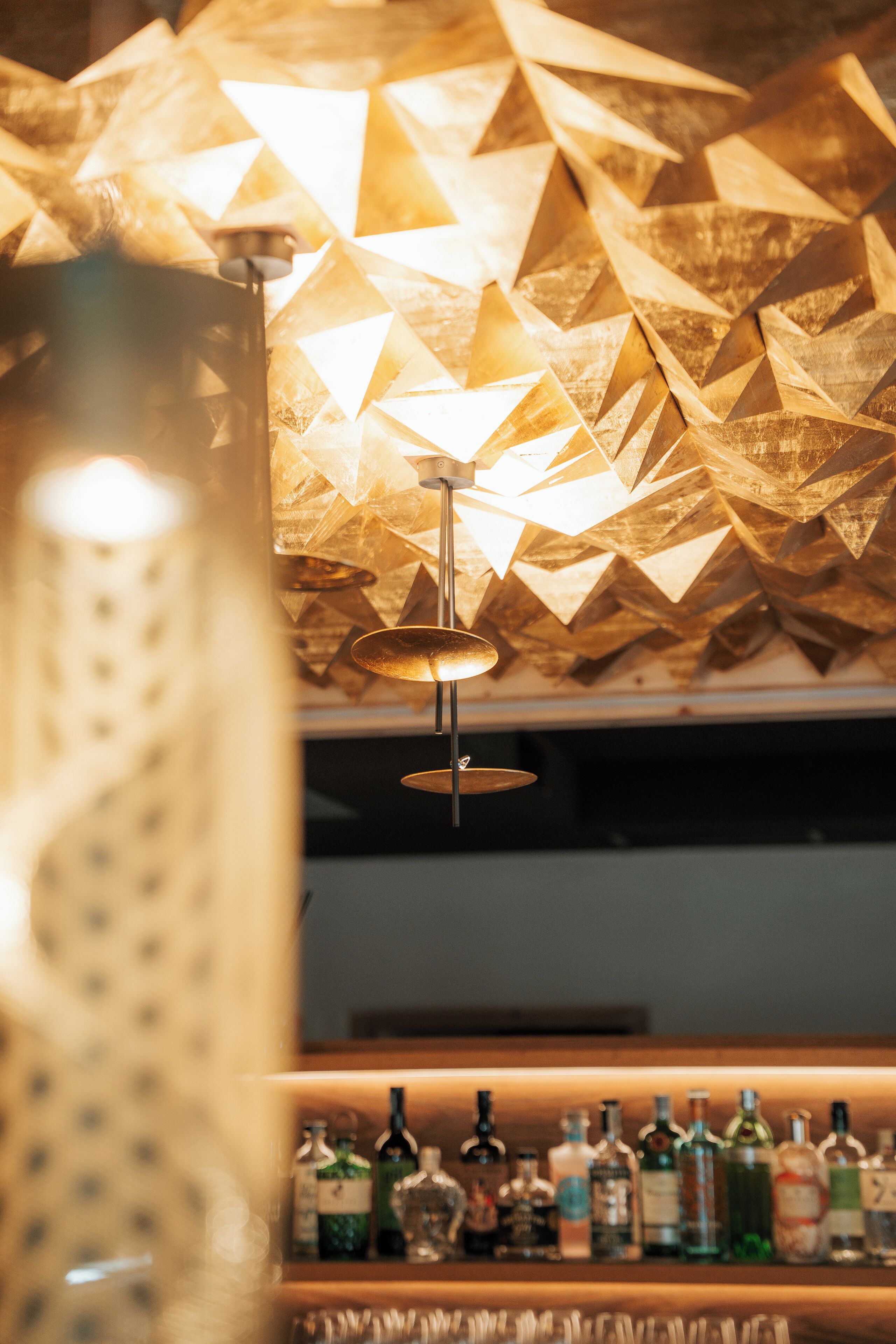 A bar area with a geometric patterned ceiling and a variety of liquor bottles on shelves.