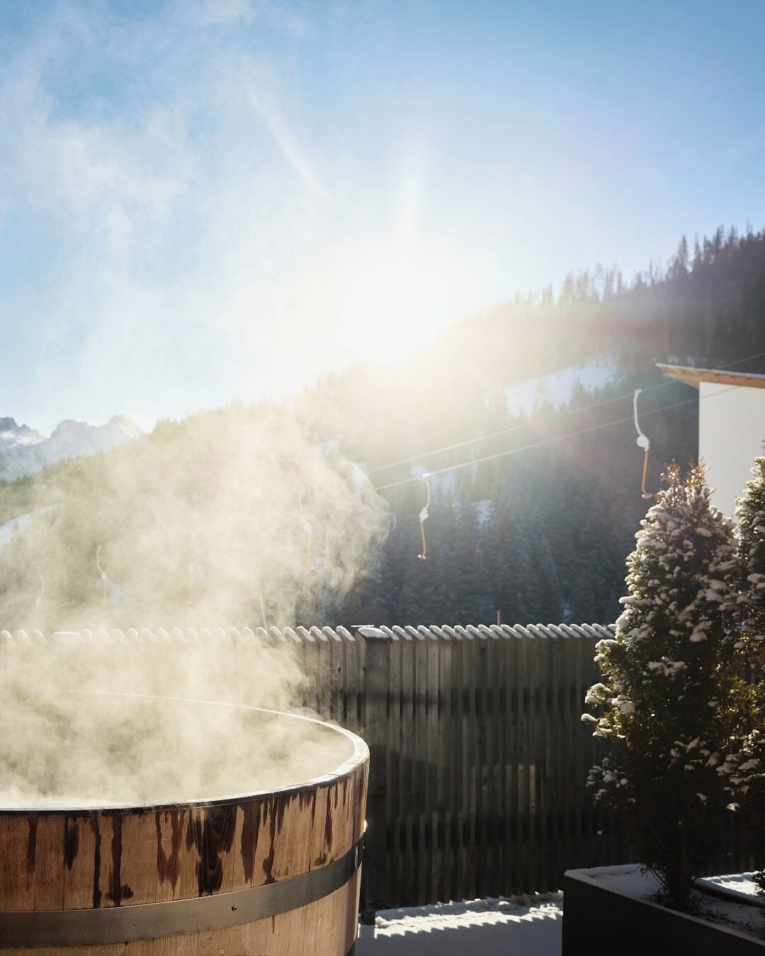 Un bain à remous dont s’échappe de la vapeur, entouré d’une clôture en bois et d’un arbre recouvert de neige.