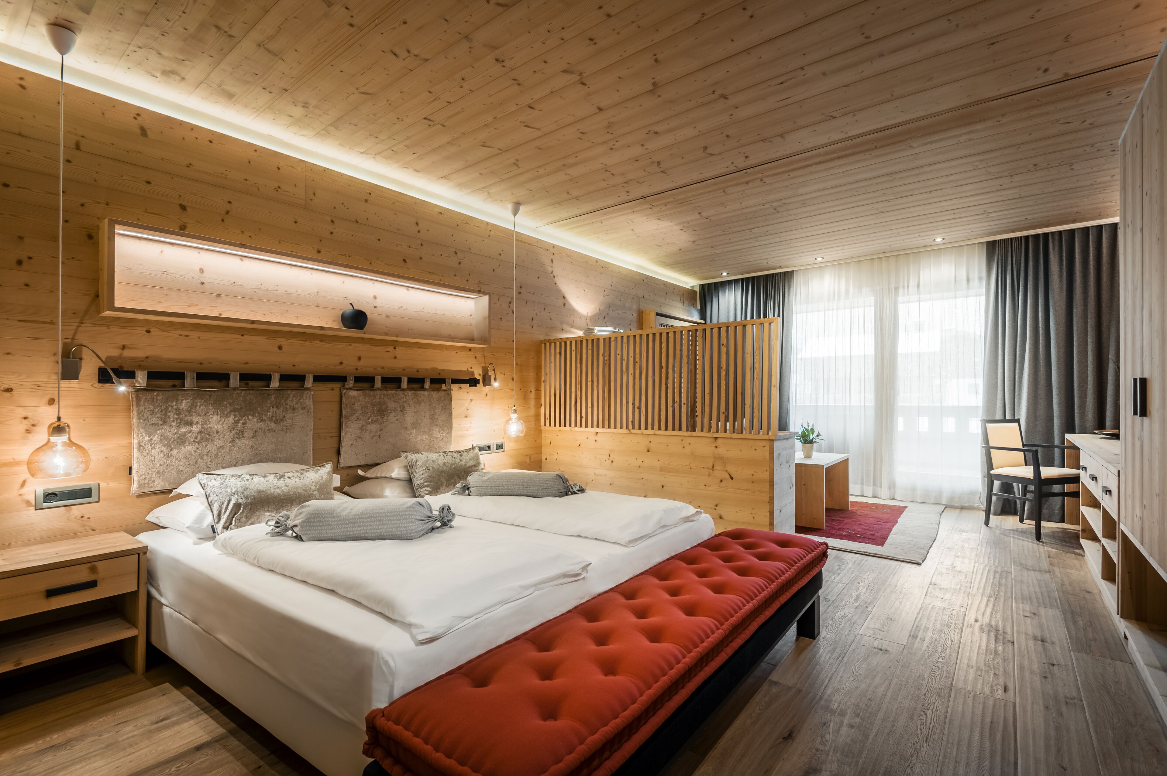 A modern bedroom with a wooden ceiling, a large bed with white linens, a red bench, and a desk with a chair.